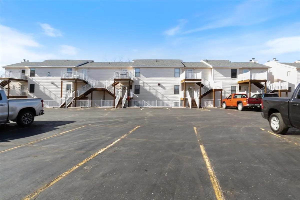 An exterior shot of the two-story apartment building, featuring staircases leading to entrances and parked vehicles. The image suggests a communal atmosphere, with balconies that hint at outdoor gatherings and relaxation spaces.