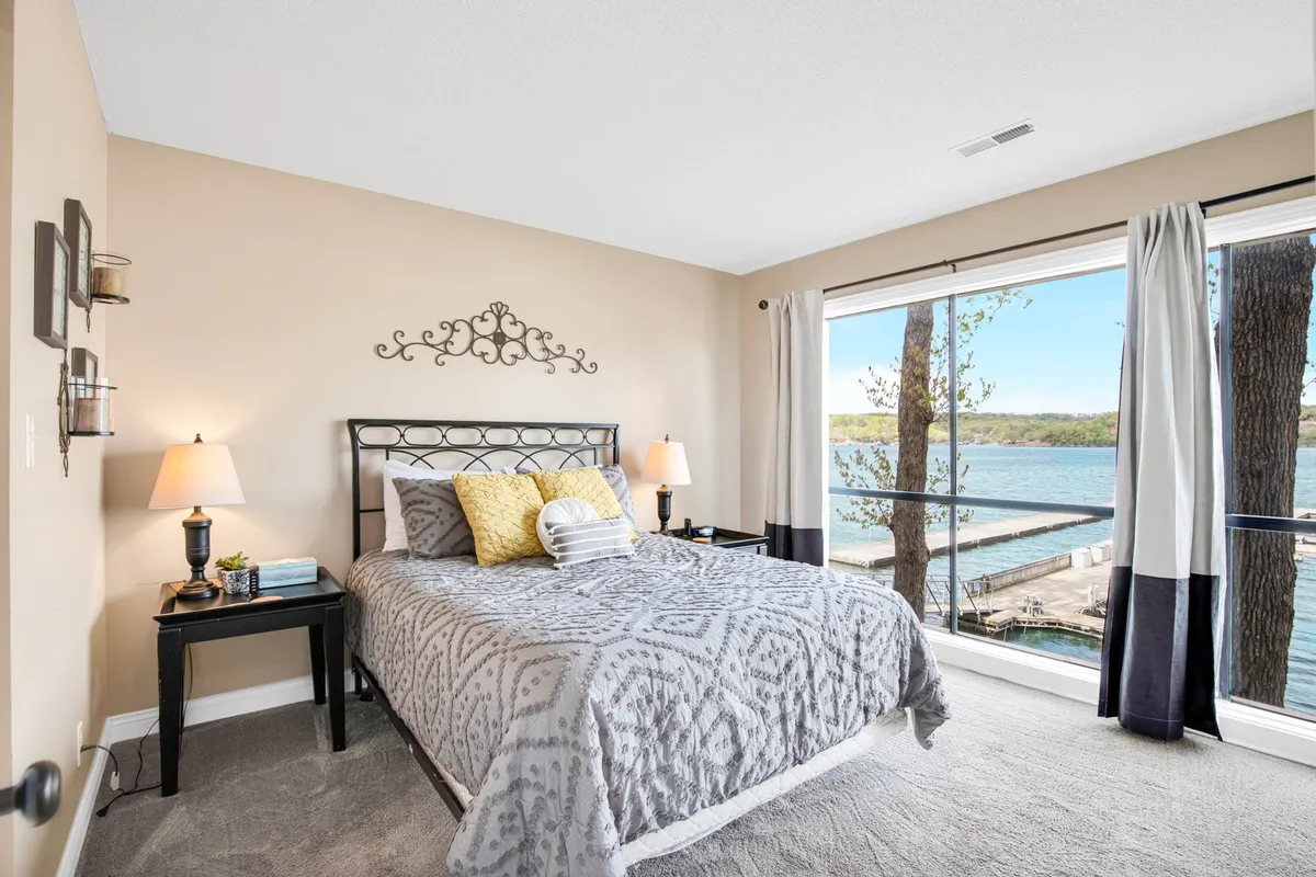 A serene bedroom with a generous window overlooking the lake, a neatly made bed, decorative wall art, and nightstands. The setting is peaceful and inviting, ideal for relaxation and enjoying the morning sunlight.