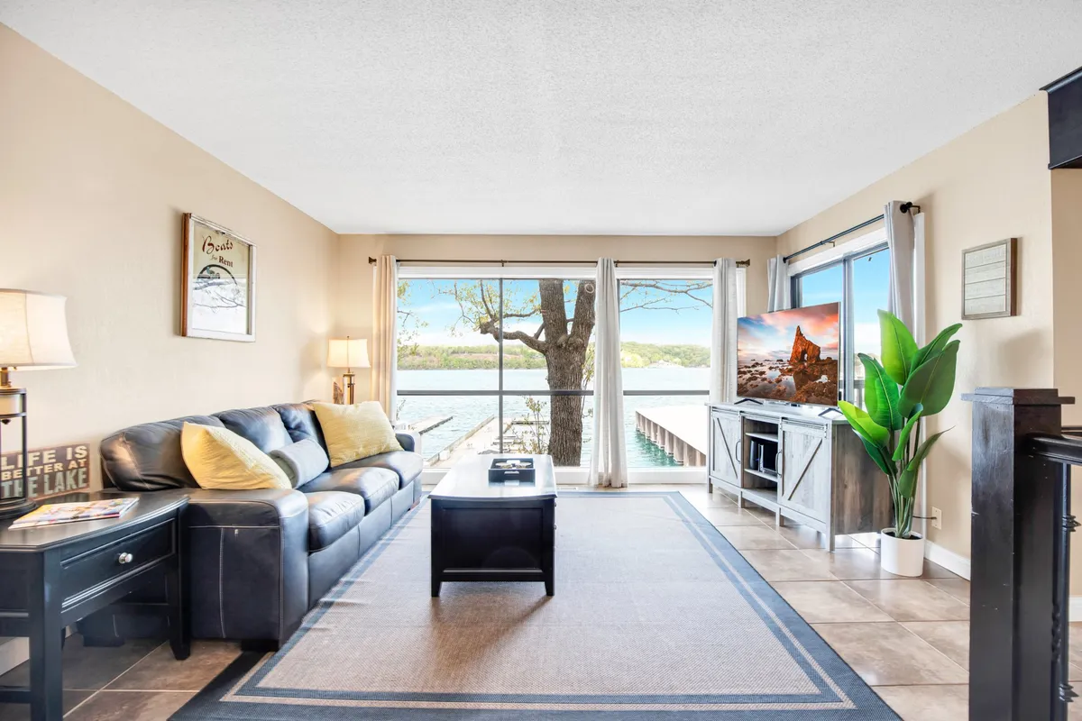 A cozy living room with a plush sofa facing large windows that overlook a serene lake. A television is mounted nearby, and a potted plant adds a touch of greenery, enhancing the sense of comfort and connection to nature. The setting bathes in warm, natural light, promising sunset views over the water.