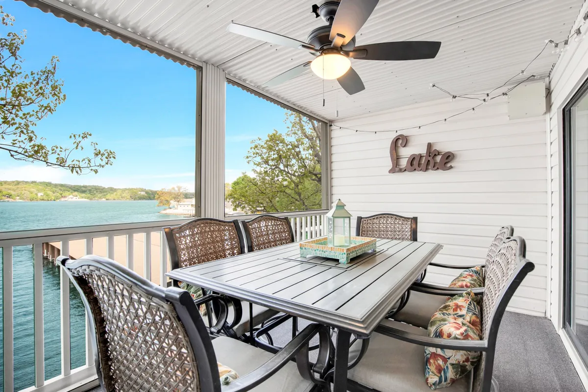 The image shows an inviting outdoor dining area on a private balcony, featuring a stylish dining table with a decorative lantern centerpiece and a ceiling fan above for comfort. Beyond, sweeping views of Lake Ozark stretch out, offering a picturesque backdrop for meals or evening gatherings. The scene evokes a sense of serenity and togetherness, perfectly suited for family meals, celebrations, or simply soaking in the lakeside sunset.