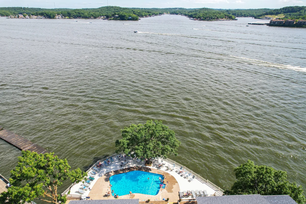 An inviting scene of a lakeside swimming pool, with people enjoying the water and a dock that extends out into the lake. The image connects the pool, dock, and open lake, all surrounded by greenery and a relaxed atmosphere.