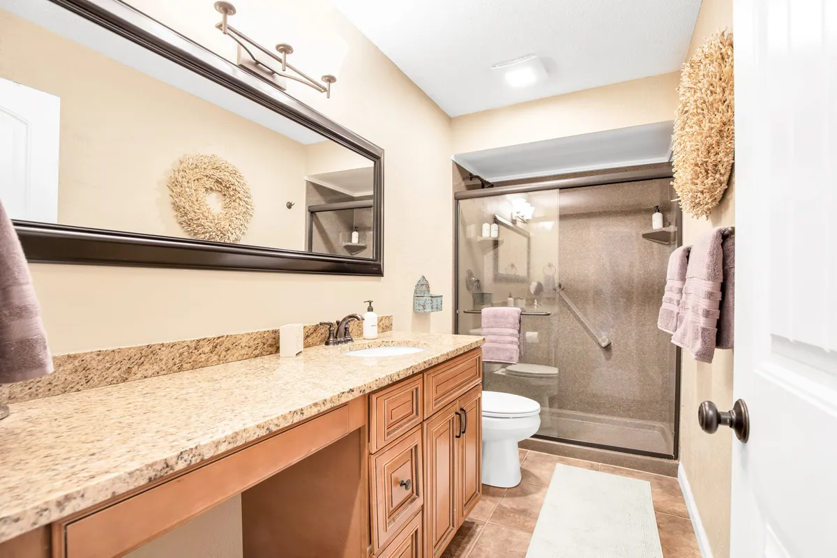 A modern bathroom with a sleek granite countertop, large mirror, stylish shower area, and decorative wall wreath. The atmosphere is calm and inviting, perfect for relaxation and self-care routines.