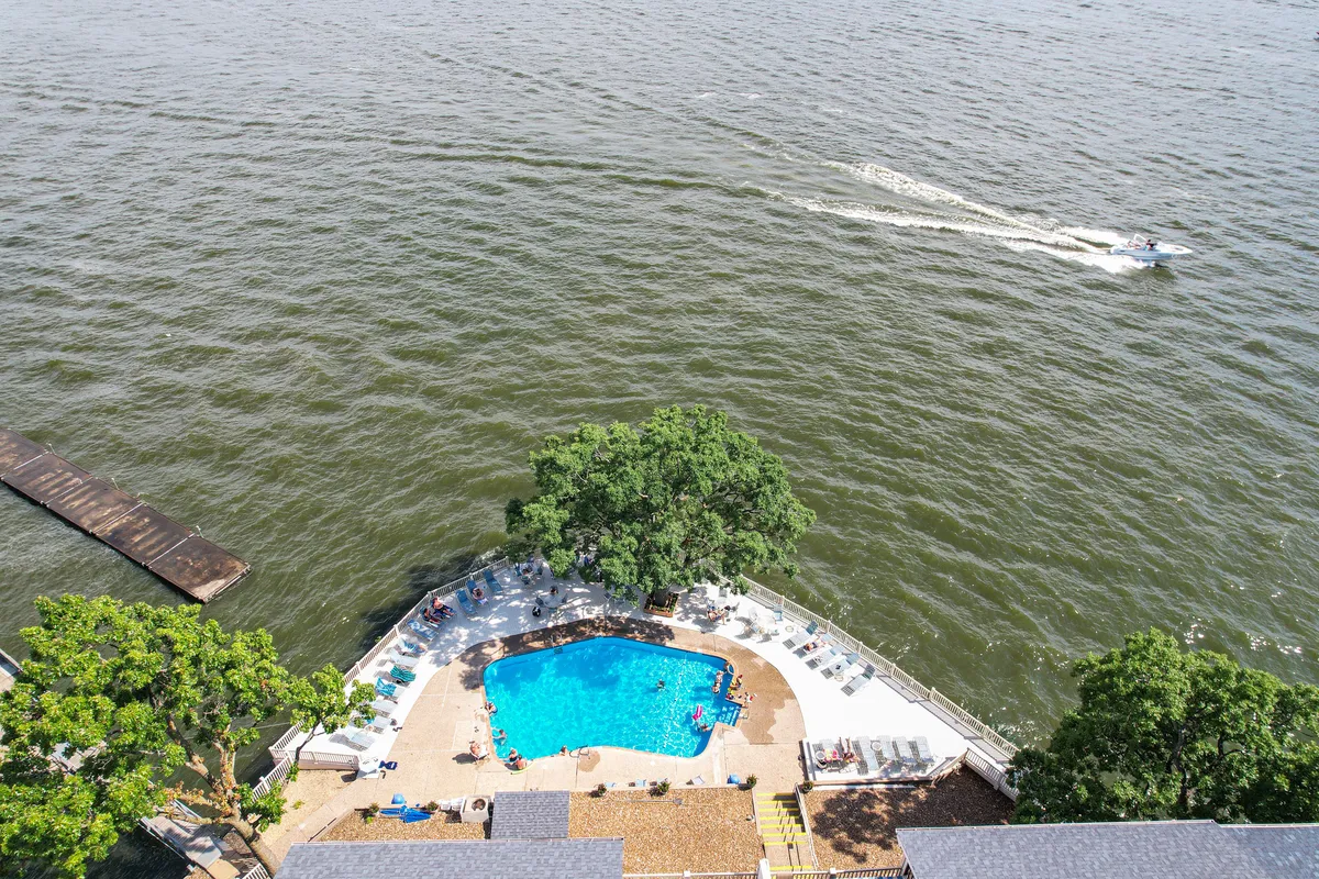 A sunlit swimming pool sits prominently in the foreground, surrounded by leafy trees and the shimmering lake beyond. A dock extends into the water, evoking possibilities for boating, paddling, or quiet reflection. The lush natural surroundings set a peaceful tone for relaxation and exploration.