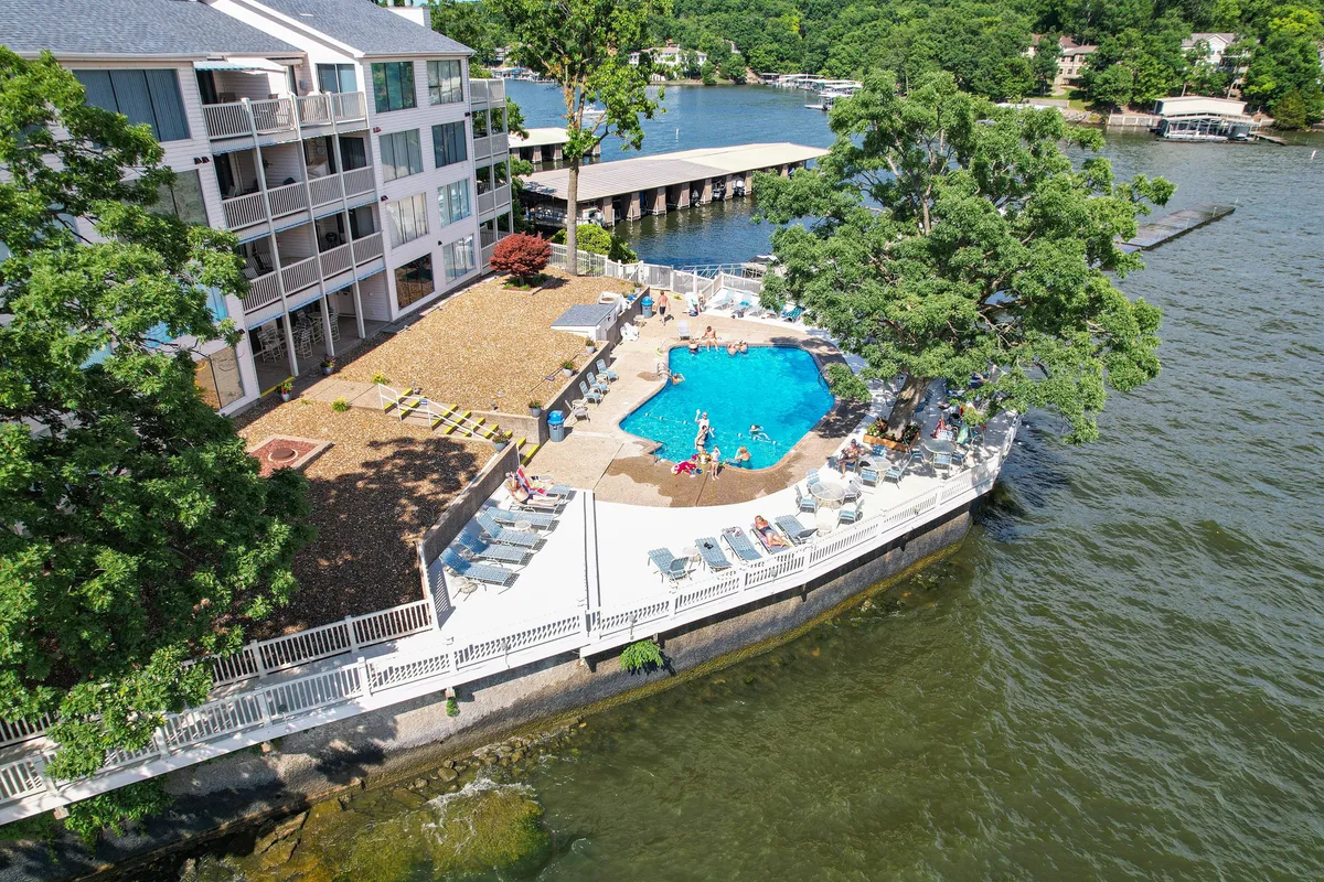 A vibrant outdoor swimming pool area framed by lush trees and a spacious deck, with the waterfront and residential building visible beyond. The scene is lively and inviting, perfect for swimming, sunbathing, and socializing with family and friends.