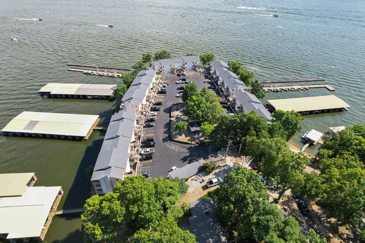 A broad exterior view featuring the waterfront, marina, docks, and picnic areas framed by trees, with residential buildings visible in the background. The scene captures the social and recreational energy of the resort surroundings.