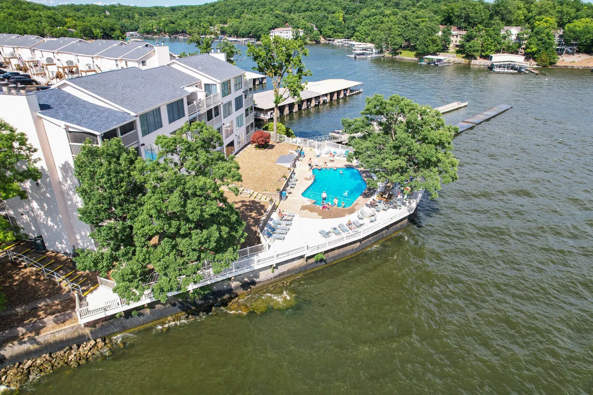 A vibrant exterior view featuring a sparkling swimming pool surrounded by lush greenery, with the lake and dock area visible in the background. People are swimming, sunbathing, and socializing poolside, with hints of boating activity nearby. The mood suggests relaxation and fun, with the amenities seamlessly blending into the lakeside setting.