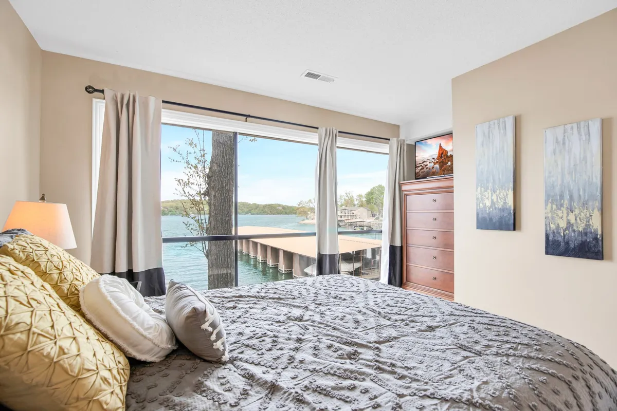 A tranquil bedroom featuring a large window framing a stunning lake view, a bed with decorative pillows, a chest of drawers, and wall art. The soft lighting and serene color palette create an atmosphere of relaxation and comfort, emphasizing the room’s peaceful retreat vibe.