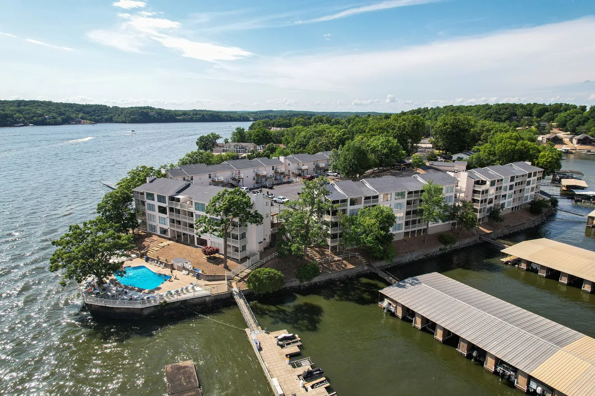 A broad exterior shot showing the swimming pool, dock, lake, and surrounding greenery, with residential buildings in the background. Various groups are sunbathing, swimming, boating, fishing, and enjoying picnics, creating a relaxed yet active lakeside ambiance.
